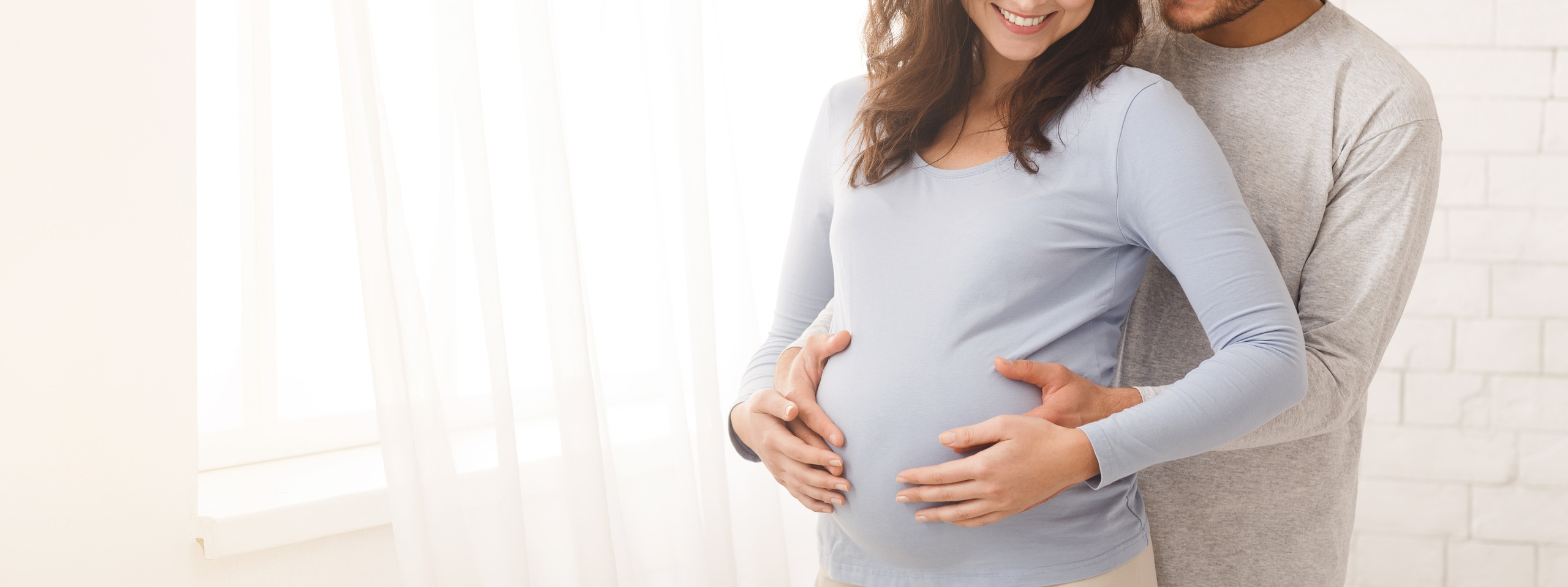 Happy pregnant couple hugging in front of the window at home.